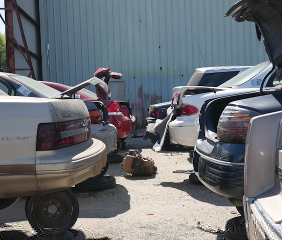 A row of cars is parked closely together in a lot, with some appearing to be missing tires, indicating a scrapyard or salvage yard setting. The cars are positioned in front of a large, corrugated metal building. Some trunks are open, and a transmission or automotive part lies on the ground.